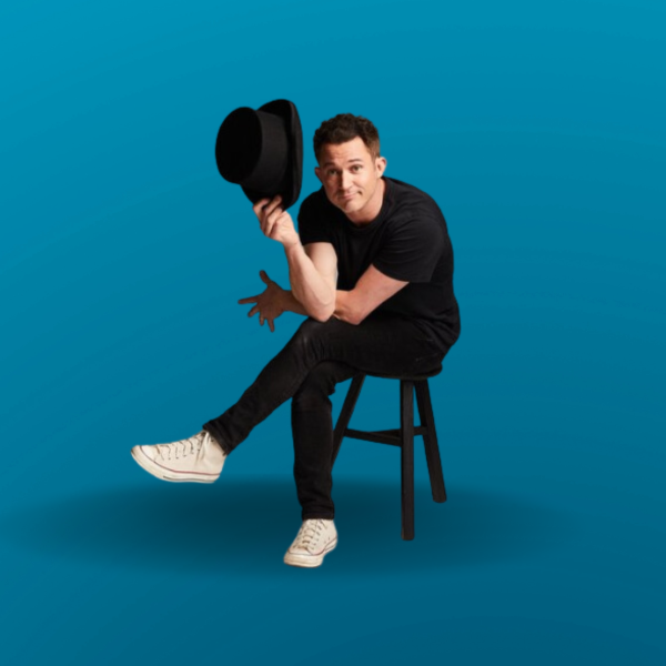 Image of magician Justin Willman sitting on a black stool and holding a black top hat. 