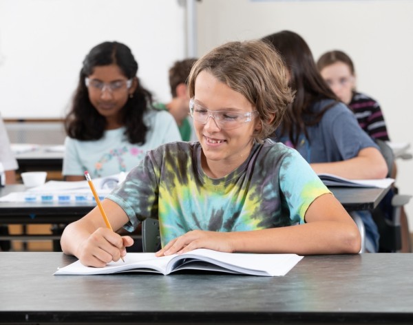 Student wearing goggles writes in a notebook in science classroom